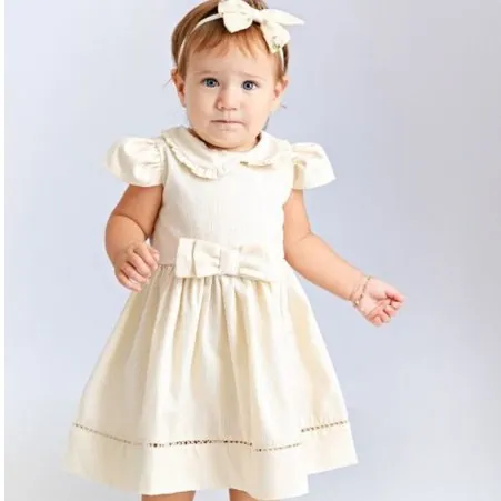 Bebê de pé com vestido creme de gola arredondada, mangas bufantes, laço na cintura e faixa de renda perto da barra