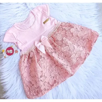 Vestido de bebê com blusa rosa-claro, cintura de pérolas, laço e saia de renda rosê sobre tapete branco felpudo