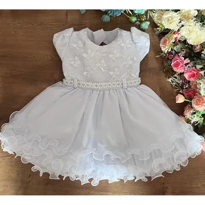 Vestido de bebê branco estendido sobre madeira, com corpo bordado de borboletas, cinto de pérolas e saia de tule ondulada