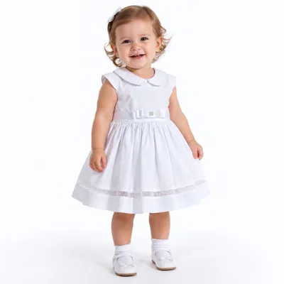 Bebê sorridente de pé com vestido branco de golinha redonda, faixa com laço na cintura, meias e sapatos brancos