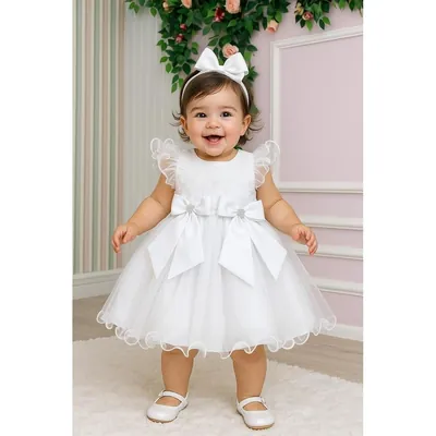 Bebê em pé com vestido branco de tule, manguinhas de babado e dois laços grandes na cintura