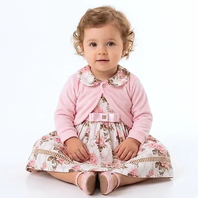 Bebê sentada com vestido estampado de rosas, gola boneca, laço rosa na cintura e bolero rosa claro