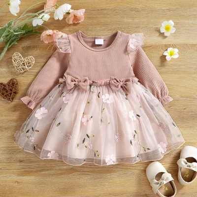 Vestido infantil rosé de manga comprida, corpo canelado, dois laços e saia de tule bordada sobre piso de madeira