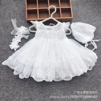 Vestido de bebê branco de renda com saia de tule em camadas, em cabide de pérolas, ao lado de touca e faixa de flores