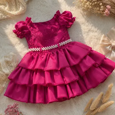 Vestido de bebê rosa-pink com mangas bufantes, flores no corpo, cinto de pérolas e saia de três babados sobre tapete claro