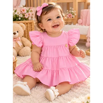 Bebê sorridente com vestido rosa de mangas de babado e saia em camadas, laço rosa no cabelo e sapatinhos brancos