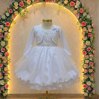 Vestido infantil branco com bolero de manga longa, cinto de pérolas e saia de tule em manequim diante de arco de rosas