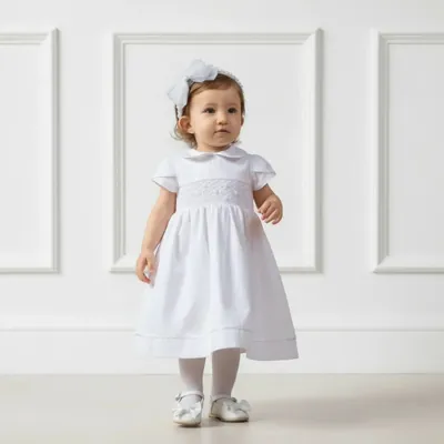 Bebê de pé com vestido branco de gola boneca, faixa bordada no peito e barra com viés, tiara de laço e sapatilhas