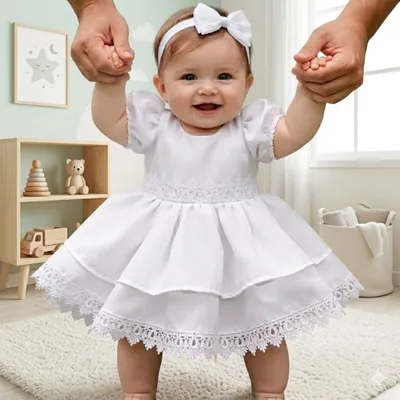Bebê sorridente de vestido branco com mangas bufantes, saia em dois babados com barra de renda e tiara com laço