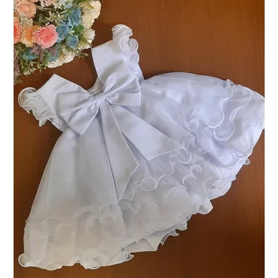 Vestido branco de bebê visto de costas, com laço grande acetinado e saia de babados ondulados, sobre mesa de madeira