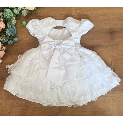 Vestido branco infantil visto de costas sobre mesa de madeira, com laço, manga bufante e saia de tule com babados ondulados