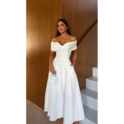 Mulher com vestido branco midi ombro a ombro, decote dobrado em laço, saia evasê e bolsa em forma de rosa, ao lado de escada