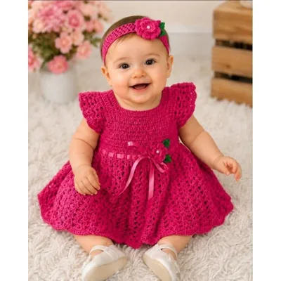 Bebê sentada em tapete branco usando vestido de crochê pink com fita e flor, e tiara de crochê com flor na cabeça