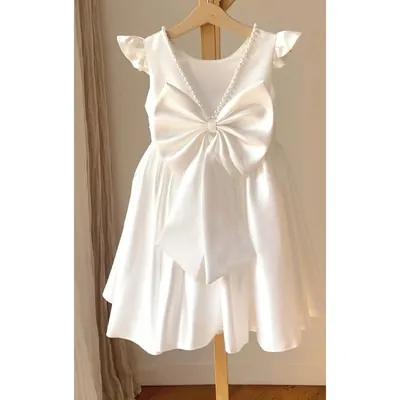 Vestido infantil branco acetinado visto de costas, com laço grande na cintura, pérolas no decote e manga de babado