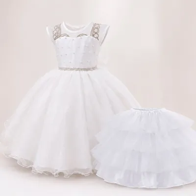 Vestido infantil branco com perolinhas, cinto de strass e saia de tule, ao lado de um saiote de babados