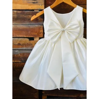 Vestido infantil off-white sem manga visto de costas, com decote em V e laço grande, pendurado em cabide de madeira