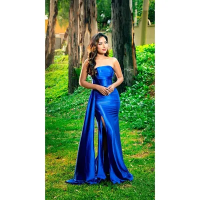 Mulher em jardim com árvores usando vestido longo azul royal tomara que caia, brilhante, com fenda e faixa lateral até o chão