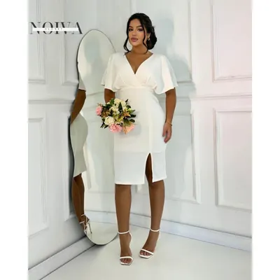 Mulher com vestido midi off-white de decote em V, mangas amplas e fenda frontal, segurando buquê ao lado de espelho oval