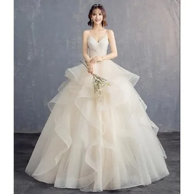 Noiva com vestido champanhe de alças finas, decote em V e saia longa de tule com babados em camadas, segurando buquê