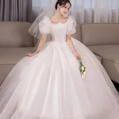 Noiva sentada em sofá bege com vestido branco de mangas bufantes, saia ampla de tule, véu curto e tulipas rosadas