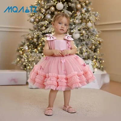 Menina pequena de vestido rosa com laços nos ombros, borboletas e saia de tule em babados diante de árvore de Natal