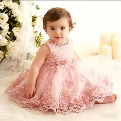 Bebê sentada descalça com vestido rosê de renda, gola de pérolas e saia de tule bordada, entre flores brancas e velas