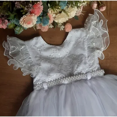 Vestido infantil branco bordado com mangas de babado transparente, cinto perolado e saia de tule, ao lado de flores