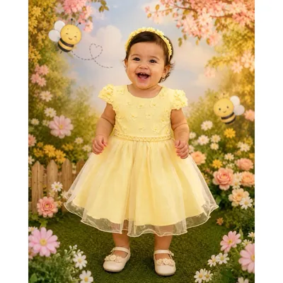 Bebê sorrindo com vestido amarelo de flores em relevo, cinto de pérolas e saia de tule, em cenário de jardim com abelhas