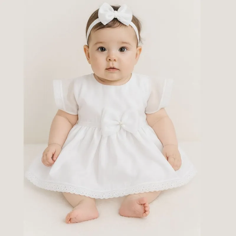 Bebê sentada com vestido branco de mangas transparentes, laço na cintura, barra de renda e tiara com laço branco