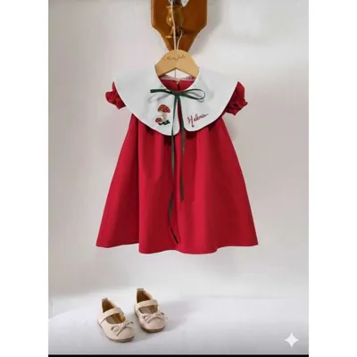 Vestido infantil vermelho com gola branca bordada com cogumelo e nome, laço verde, pendurado em cabide de madeira
