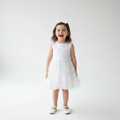 Menina sorrindo com vestido branco sem mangas, corpo liso e saia de renda bordada, laço branco no cabelo e sapatilhas
