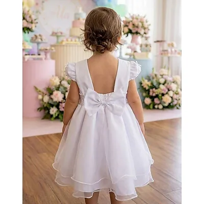 Menina de costas com vestido branco de saia em camadas, decote nas costas e laço grande, em salão decorado com flores