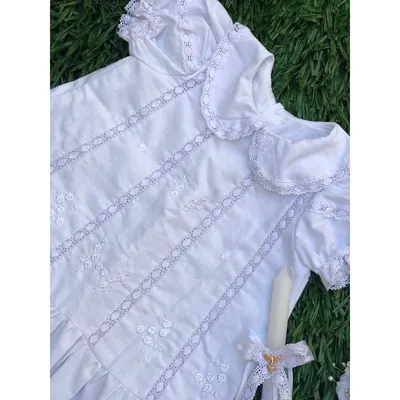 Vestido infantil branco sobre a grama, com gola boneca rendada, faixas verticais de renda, flores bordadas e vela ao lado