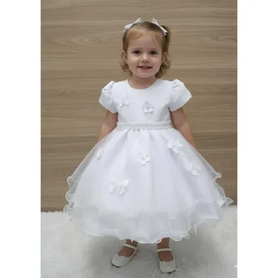 Menina pequena de pé usando vestido branco com mangas bufantes, flores aplicadas, cinto de pérolas e saia de tule ondulada