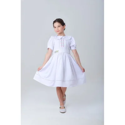Menina com vestido branco de gola de babado, flores bordadas no peito e faixa com as letras ABC na cintura