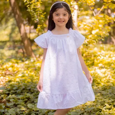 Menina sorridente com vestido branco de laise, mangas curtas franzidas e babado na barra, em um jardim ensolarado