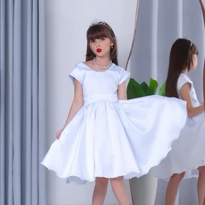 Menina de tiara usando vestido branco rodado de manguinha e faixa na cintura, refletida em espelho