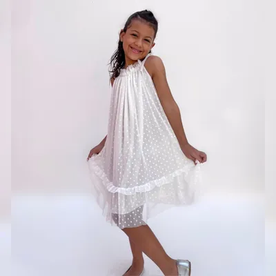 Menina sorrindo com vestido branco de tule poá, gola franzida, alças amarradas em laço, babado na barra e sapatilha prata