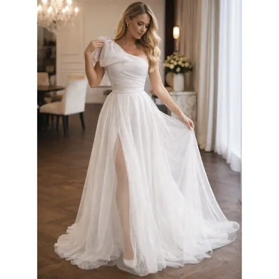 Mulher loira em salão com lustre usando vestido branco longo de um ombro, laço no ombro, saia brilhante com fenda