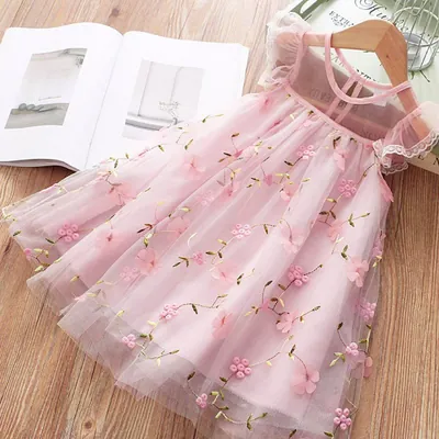 Vestido infantil rosa de tule com flores bordadas pendurado em cabide de madeira sobre mesa com revista aberta