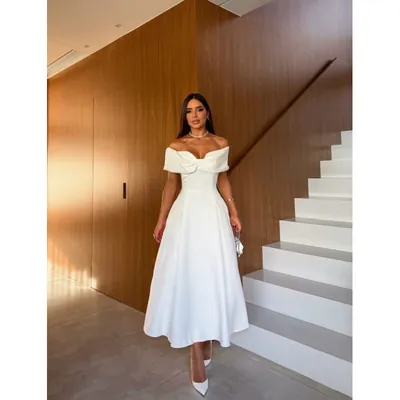 Mulher de vestido midi branco ombro a ombro com nó no decote e saia evasê, ao lado de escada branca e parede de madeira