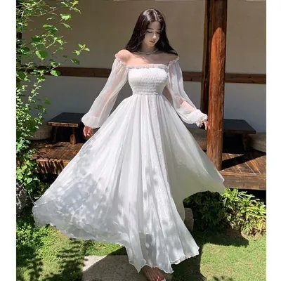 Mulher descalça num jardim girando com vestido branco longo ombro a ombro, corpete franzido e mangas bufantes transparentes