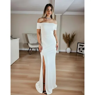 Mulher em sala clara usando vestido branco longo ombro a ombro, liso, ajustado, com fenda alta na perna direita