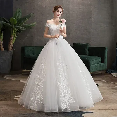 Noiva com vestido princesa ombro a ombro, saia de tule com ramos bordados e flor rosa na mão, diante de sofá verde
