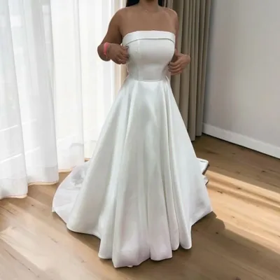 Mulher em quarto iluminado vestindo vestido de noiva branco tomara que caia, liso, com saia evasê e pequena cauda