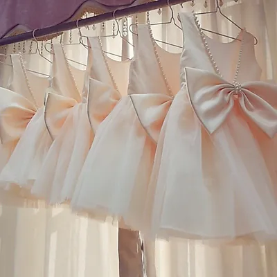 Vestidos infantis cor champanhe pendurados em cabides, vistos de costas, com laço grande, pérolas e saia de tule